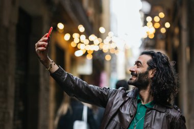 Gülümseyen turist, arka planda güzel bokeh ışıkları olan tarihi bir Avrupa şehir merkezinde selfie çekiyor.