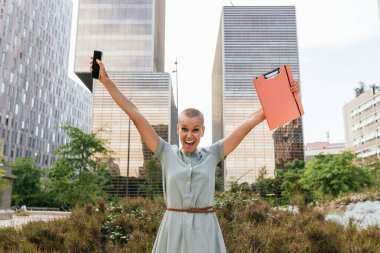 Energetic young adult businesswoman celebrating professional success and financial achievement outdoors in the modern urban cityscape. Surrounded by office buildings and skyscrapers clipart