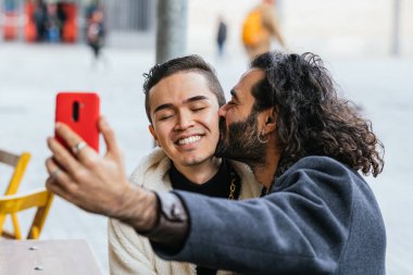 Bir selfie için öpüşen ve gülümseyen iki adam, samimi bir sokak sahnesinde aşk ve dostluk gösteriyorlar.
