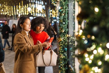 İki genç kadın noel alışverişi yaparken ve akıllı telefondan anlaşmaları kontrol ederken eğleniyorlar.