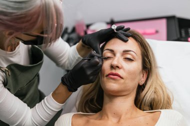 Siyah eldivenli makyaj sanatçısı, güzellik salonunda beyaz bluz giyen rahat bir kadına özel kaş estetiği uyguluyor.