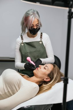 Yetenekli bir güzellik uzmanı modern, hijyenik güzellik salonundaki bir bayan müşterinin dudaklarında mikro pigmentasyon yapıyor.