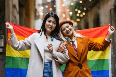 Two young women are holding a large rainbow flag and smiling while holding blue lollipops