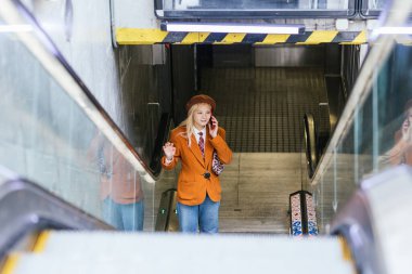 Genç iş kadını metro istasyonunda yürüyen merdivenden çıkarken telefonla konuşuyor.