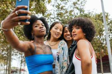 Parkta selfie çeken dört neşeli kadın arkadaşlığı, çeşitliliği ve kendini kabulü kutluyorlar.