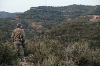 Tüfekli avcı tepenin üstünde dikilip vadiye bakarken manzaranın tadını çıkarıyor.