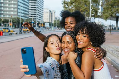 Dört neşeli kadın şehirde bir akıllı telefonla selfie çekiyor ve sarılıyorlar.