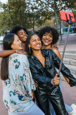 Şehir parkında selfie çubuğuyla selfie çeken dört mutlu arkadaş.