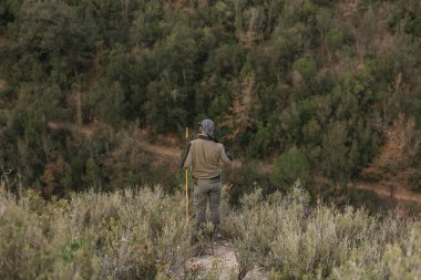 Hunter bir tepede dikiliyor, omzunda bir tüfekle uçsuz bucaksız bir ormana bakıyor.