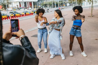 Arkadaşları şehir caddesinde akıllı bir telefonla video çekerken üç kız dans edip poz veriyor.