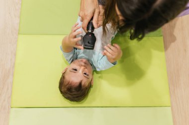 Optometrist is using a retinoscope during an eye exam on a baby lying on a green mat clipart