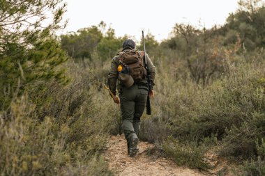 Avcı ormanda yürüyor. Toprak bir patikada. Tüfek ve sırt çantası taşıyor.
