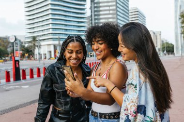 Üç neşeli genç kadın, canlı bir şehir ortamında akıllı telefondan eğlenceli bir şeye bakarken birlikte bir anlık kahkaha atıyorlar.