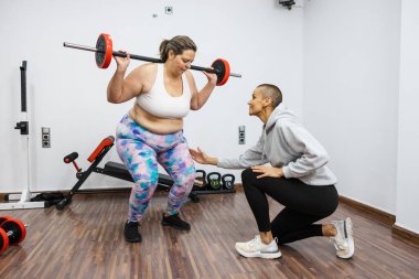 Kilolu bir kadın kişisel antrenörüyle ağırlık kaldırıyor spor salonunda yardımcı oluyor, sağlıklı yaşam tarzını ve formda kalmayı teşvik ediyor.