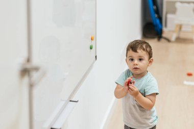 Toddler standing next to an eye chart, playing with a toy during a pediatric optometry checkup clipart