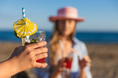 Güneşli sahilde ferahlatıcı limon dilimi, nane ve şemsiye ile el ele tutuşmak ve arka planda bulanık bir kadın.