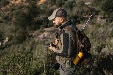 Avcı ormanda tüfek ve sırt çantası taşırken telsiz kullanarak kamuflaj kıyafeti giyiyor.