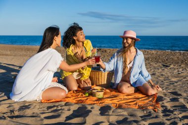 Sahilde piknik yapan üç farklı kadın güneşli bir günde serinletici kokteyllerle kadeh kaldırıyor.