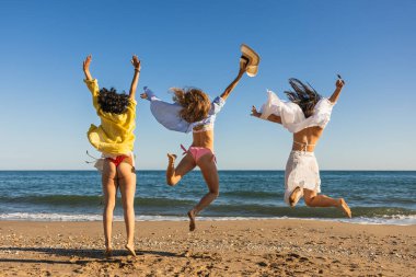 Üç neşeli çoklu etnik kadın sahilde zıplıyor, güneşin tadını çıkarıyor ve yaz tatillerinde eğleniyorlar.