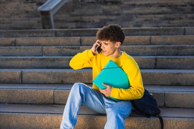 Genç bir erkek öğrenci kolej binasının merdivenlerinde otururken elinde bir klasör ve akıllı bir telefonla konuşuyor.