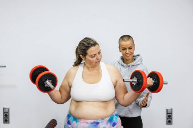 Kilolu bir kadın kişisel antrenör yardımıyla ağırlık kaldırıyor, spor salonundaki fitness hedefleri doğrultusunda birlikte çalışıyor.