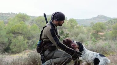 Deneyimli avcı başarılı bir yolculuktan sonra iki sadık av köpeğini nazikçe okşadı, engebeli dağ manzarası arasında derin bir bağ olduğunu gösterdi.