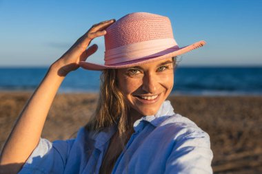 Gün batımında kumsalda pembe bir şapkayla gülümseyen beyaz bir kadın okyanusun sıcacık altın saatinin tadını çıkarıyor.