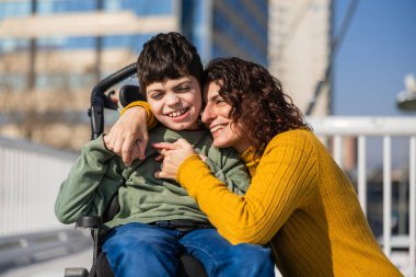 Serebral felçle oğlunu kucaklayan mutlu bir anne güneşli bir günde dışarıda tekerlekli sandalyede oturuyor.