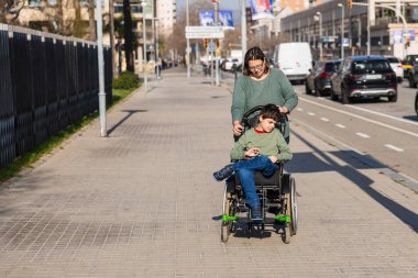 Şefkatli bir anne oğlunu engelli bir halde şehir kaldırımında tekerlekli sandalyeye itiyor. Destek ve hareketlilik sağlıyor.