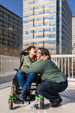 Sevgi dolu anne, neşeli oğlunu beyin felciyle kucaklıyor, şehir merkezinde bir köprüde birlikte vakit geçiriyorlar.