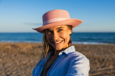 Beyaz kadın kumsalda gülümsüyor pembe şapka ve mavi gömlek giyiyor, yaz tatilinin tadını çıkarıyor.
