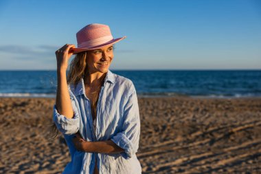 Mutlu kadın pembe şapkasını tutarken gülümsüyor, şık mavi bir gömlek giyiyor, güzel bir sahilde güneşin tadını çıkarıyor.