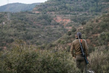 Hunter ormanda duruyor, omzuna bir tüfek dayayıp, uzaklığa bakıyor.