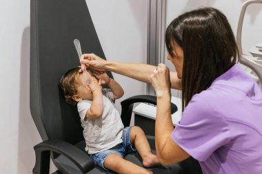 Optometrist, görme testi sırasında sandalyede oturan bir çocuğun bir gözünü kapatıyor.