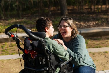 Mutlu anne engelli oğluyla oynuyor. Parkta tekerlekli sandalyede oturuyor, birlikte vakit geçiriyor.
