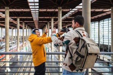 İki erkek turist, hareketli bir tren istasyonunda macera ve yaratıcılığı kucaklayarak eğlenirken komik videolar ve fotoğraflar çekiyor.