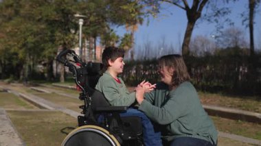 Sevgi dolu bir anne oğluyla sıcak bir ilişki içinde, tekerlekli sandalyede beyin felci geçiriyor, açık hava parkında eğlenceli anlar yaşıyor, aile bağlarını ve destekleyici bakımı gösteriyor.