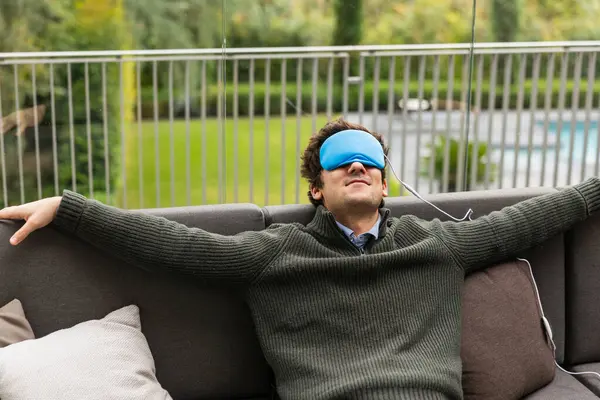 Vücut ısısı kontrollü uyku maskesi takan bir adam koltukta kollarını açmış bir şekilde rahatlar ve bir anlık huzurun tadını çıkarır.