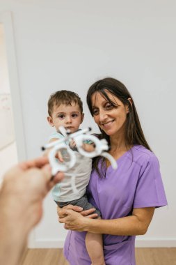 Bir göz doktoru görme testi sırasında çocuğa bir deneme çerçevesi gösterirken gülümseyen pediatrik optometrist kucağında bir bebek tutuyor.
