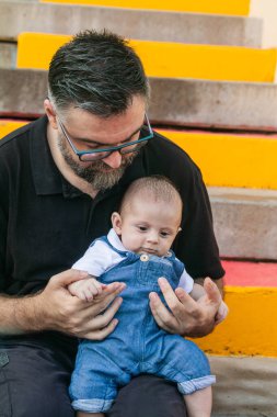 Baba dört aylık bebeğini kucağında canlı, renkli merdivenlerde otururken dışarıda sevgi ve bağlantının tadını çıkarıyor.