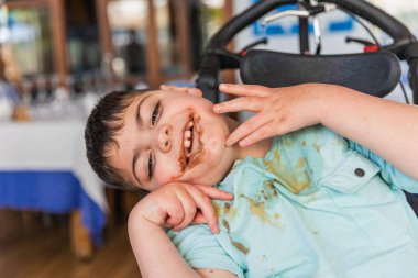 Serebral felçli genç, adaptasyon koltuğunda oturuyor, dağınık yemeğin tadını çıkarıyor ve gülümsüyor.