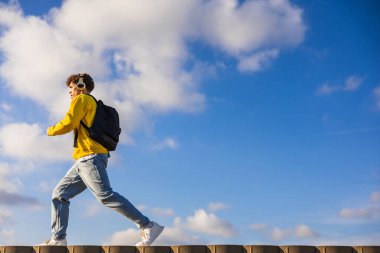 Kulaklık takan ve sırt çantası takan genç bir adam bulutlu mavi gökyüzüne karşı yürüyen güneşli bir günde müziğin keyfini çıkaran bir duvarda yürüyor.