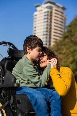 Sevgi dolu bir anne, engelli bir şekilde oğlunu kucaklıyor tekerlekli sandalyede oturuyor, açık havada sevgi ve destek anı paylaşıyor.