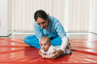 Beş aylık bir bebeği destekleyen fizyoterapist, kırmızı hasırla ilgili ilk değerlendirme sırasında kafa kontrolü ve yatkınlık eğitimi alıyor.