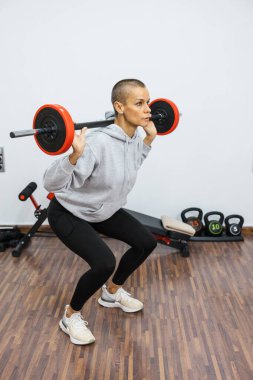 Güçlü bir kadın omuzlarında halterle çömeliyor, spor salonunda egzersiz yapıyor, dayanıklılık ve zindelik gösteriyor.