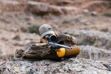 Hunter vahşi doğada tüfekle nişan alıyor. Sırt çantası bir kayanın üzerinde duruyor.