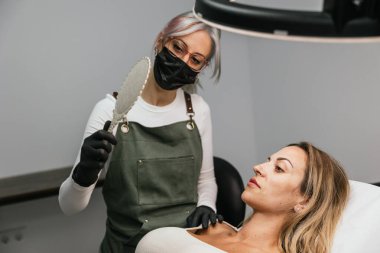 Kadın yeni mikrobıçaklı kaşlarını aynada inceliyor. Güzellik uzmanı modern bir güzellik salonunda geribildirim bekliyor.