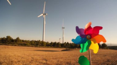 Arka planda rüzgar türbinleriyle dönen renkli fırıldak yavaş hareket ederek yenilenebilir enerjiyi sembolize ediyor.