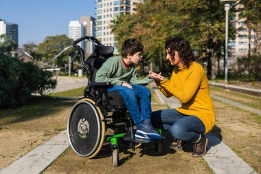 Anne, güneşli bir şehir parkında tekerlekli sandalyede oturan engelli oğluyla mutlu bir şekilde iletişim kuruyor.