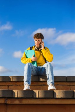 Dosyayı tutarken cep telefonu kullanan erkek öğrenci, mavi gökyüzü olan merdivenlerde oturuyor.